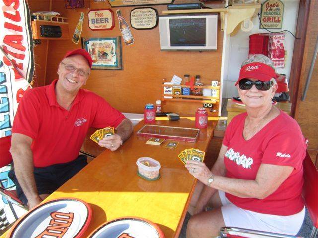 UNLV vs. Central Michigan Tailgating on September 14, 2013. Submitted by Joe Van Rhyn.
Elaine and Joe Van Rhyn tailgating at a Rebel game. Joe always enjoyed watching football in a tavern. He and Elaine have also has been 20 year season ticket holders for the Rebels. So instead of going to the tavern, he built a tavern to bring to the games. The sides and back open up, the bar and chairs come out from the storage room behind the back bar, the lights, TV and music are turned on and "the bar is open."
The bar is called "Clem's Bar" to honor Elaine's family bar that was started in Berlin, Wisconsin by her Grandfather in 1904. Except during prohibition, when it was an ice cream shop. (wink, wink) It has been a family bar ever since. Elaine's mom & dad ran it from 1941 to 1976 and her brother has run it since then. Next year the family will celebrate 110 years in business.
Joe's bar has radio, TV, a 2 burner stove, barbeque grill, pizza oven and coffee maker. It can run off battery or 110 electric. You can always find them in the Scarlet lot cooking up some interesting tailgating food. "Go Rebels!"
