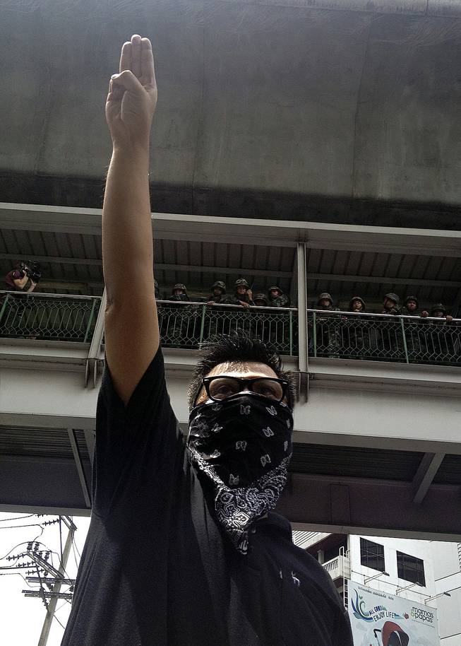 In this June 1, 2014, photo, an anti-coup protester gives a three-finger salute as soldiers keep eyes on him from an elevated walkway near a rally site in central Bangkok, Thailand. Thailand's military rulers say they are monitoring the new form of silent resistance to the coup — borrowed from "The Hunger Games" — and will arrest those in large groups who ignore warnings to lower their arms.