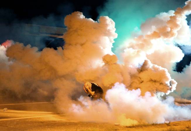 A protester takes shelter from smoke billowing around him Wednesday, Aug. 13, 2014, in Freguson, Mo. Protests in the St. Louis suburb rocked by racial unrest since a white police officer shot an unarmed black teenager to death turned violent Wednesday night, with some people lobbing Molotov cocktails and other objects at police who responded with smoke bombs and tear gas to disperse the crowd.